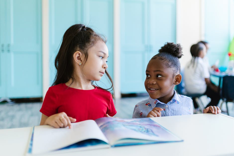 Two young children in a classroom interact while reading a book together.
