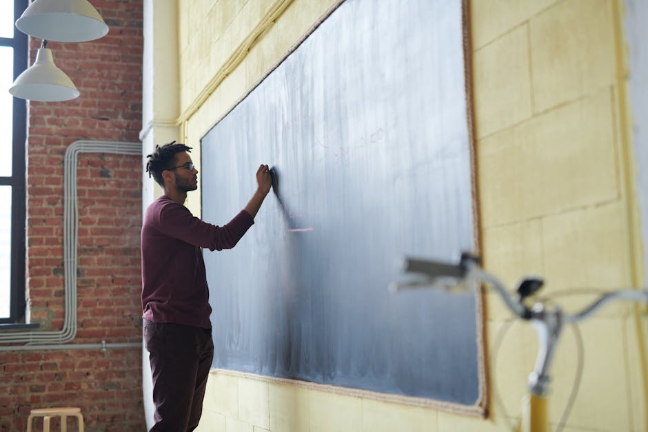 Man with dreadlocks writing on a blackboard in a classroom setting